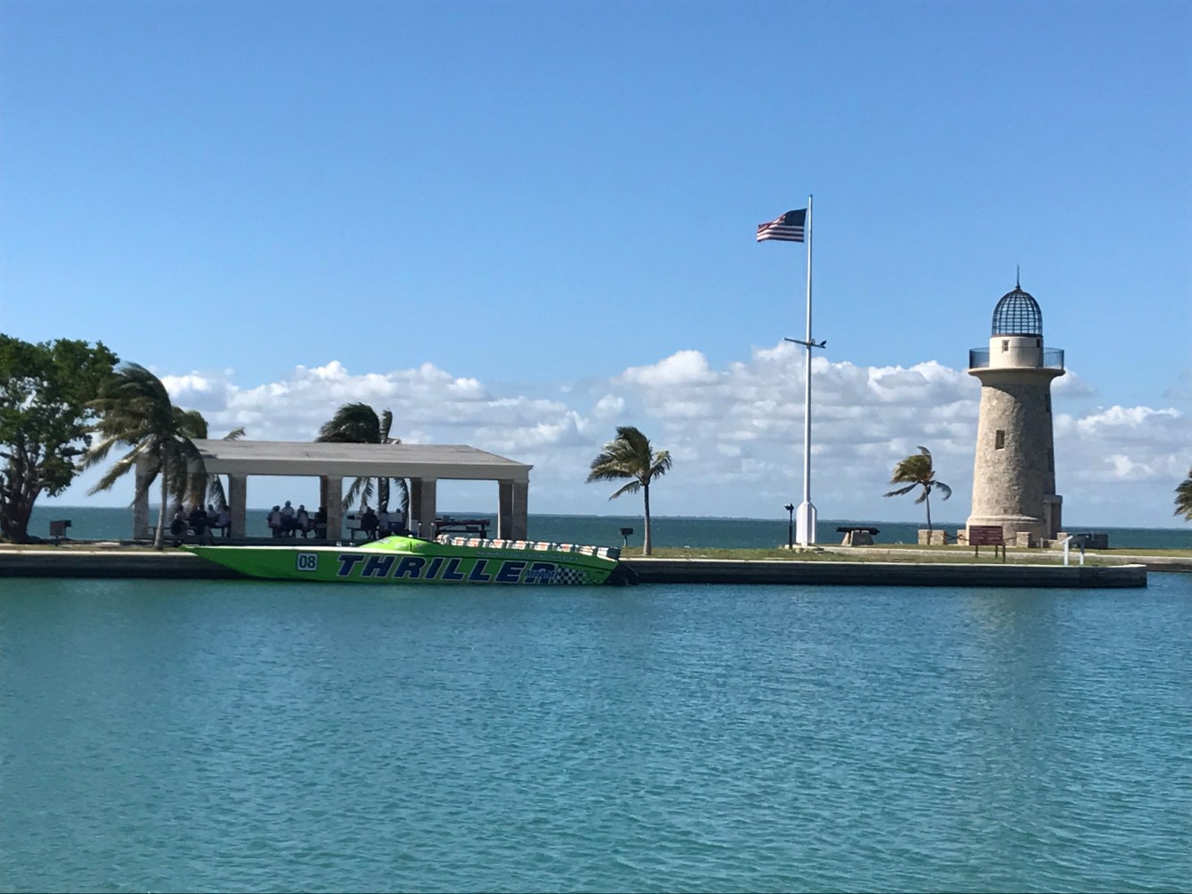 Green speedboat by pavilion and lighthouse on a sunny day with American flag, palm trees, and water.