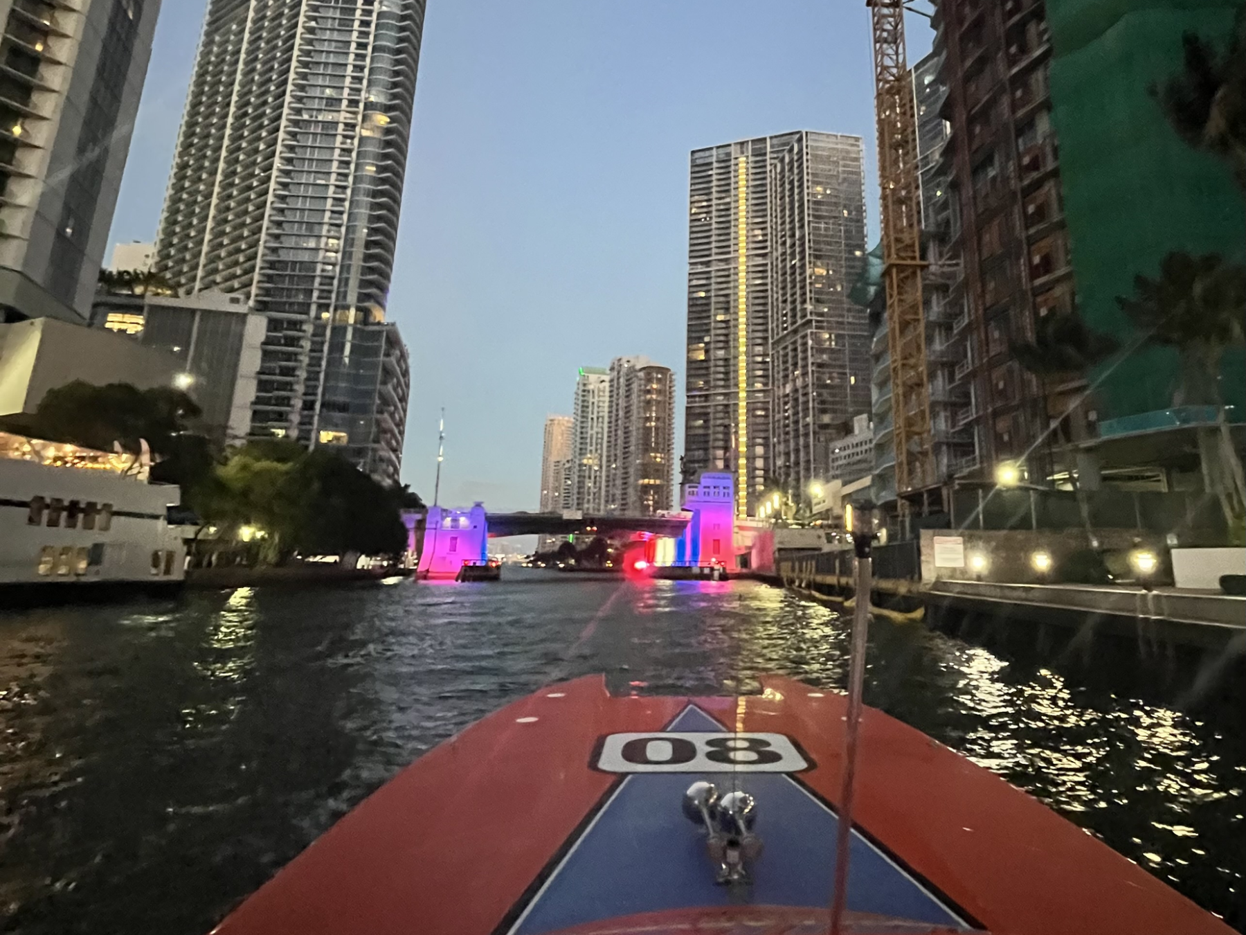 View from boat in city canal with skyscrapers and lit bridge at dusk.