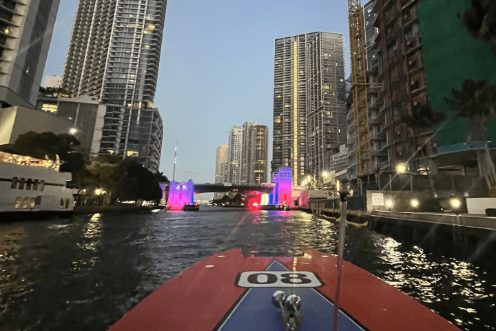 View from boat in city canal with skyscrapers and lit bridge at dusk.
