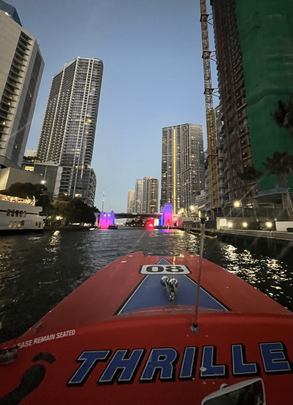 View from boat in city canal with skyscrapers and lit bridge at dusk.