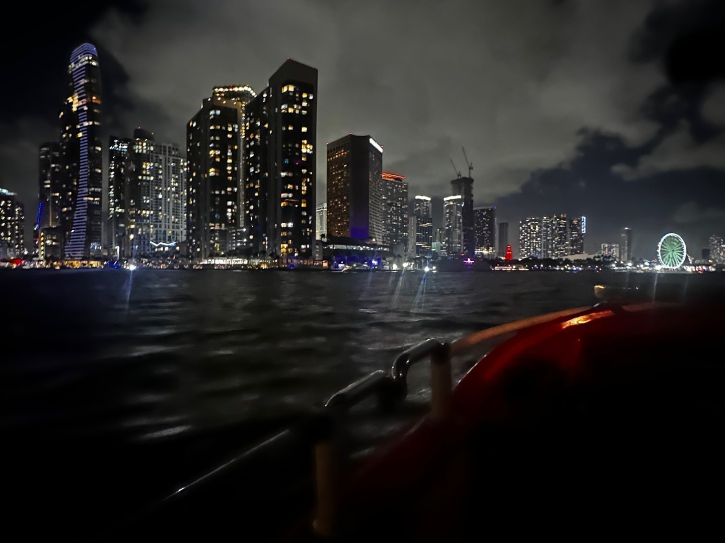 Nighttime city skyline with illuminated skyscrapers and a Ferris wheel, seen from a water perspective.