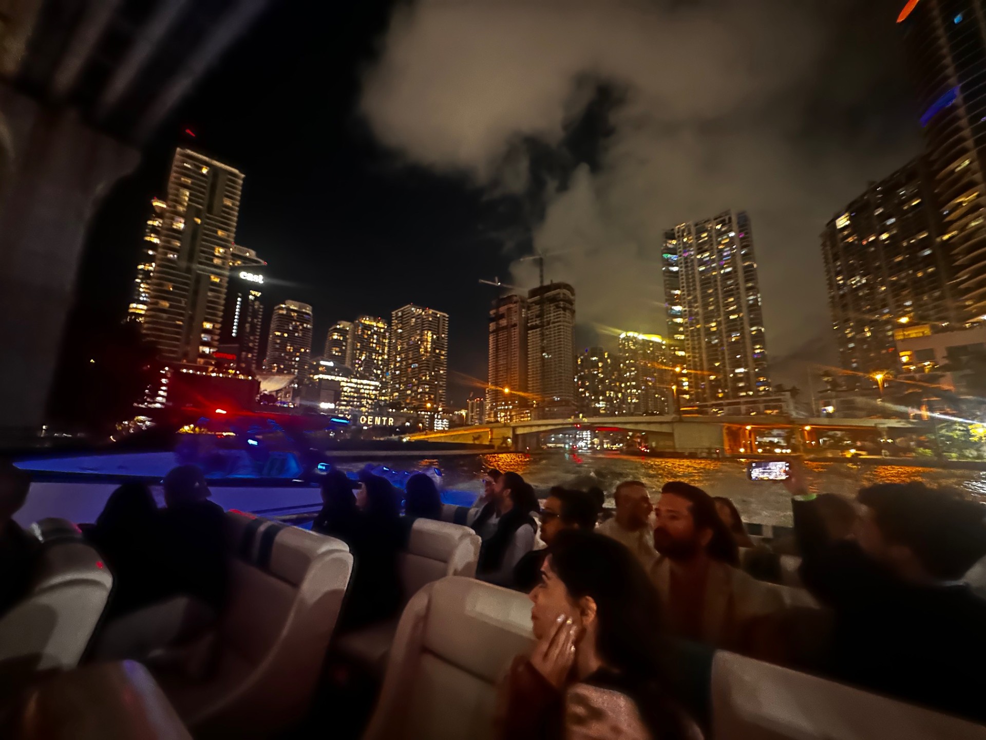 Group of people on a boat, city skyline and colorful lights at night.
